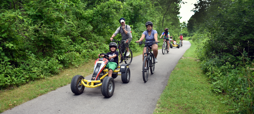 Adults riding bicycles and a child riding a four wheel pedal car on the Addison Oaks Bike Trail