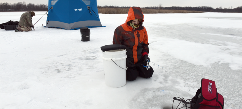 An ice fisher kneeling on the ice looking at the ice fishing hole